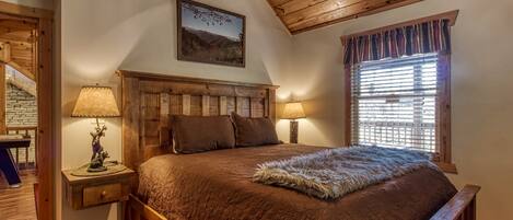 Un dormitorio con cabecera de madera, dos lámparas de noche, una cama grande con colcha marrón y una ventana con persianas.