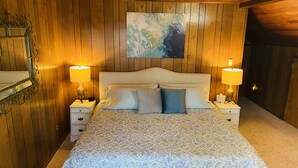 Een slaapkamer met een wand van houten panelen, een groot bed met beddengoed met een patroon, twee nachtkastjes met lampen, en een kunstwerk in een lijst boven het bed.