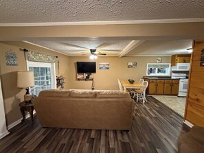 A living room with a sofa, a dining table with chairs, a television, and a kitchen area with cabinets and appliances. - Lakeside Cottage - Lake, Private Hot Tub (Guild)