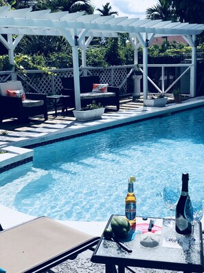 Un espace aménagé au bord de la piscine, avec une pergola, des chaises longues et une table sur laquelle se trouvent une bouteille, un verre et des fruits.