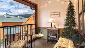 Ein gemütlicher Balkon mit Holzgeländer, zwei Stühle mit gemusterten Kissen, ein kleiner Tisch mit Lampe, ein Weihnachtsbaum und ein gerahmtes Schild an der Wand.