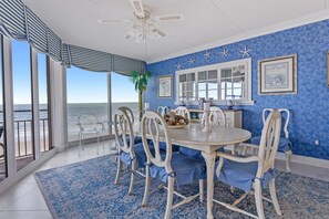 Un espace repas offrant une vue sur l’océan, avec une table ronde, des chaises blanches et un tapis à motifs bleus.