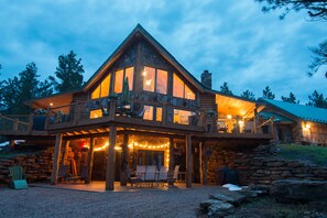 A log cabin with a large porch, stone walls, and a balcony overlooking a forested area.