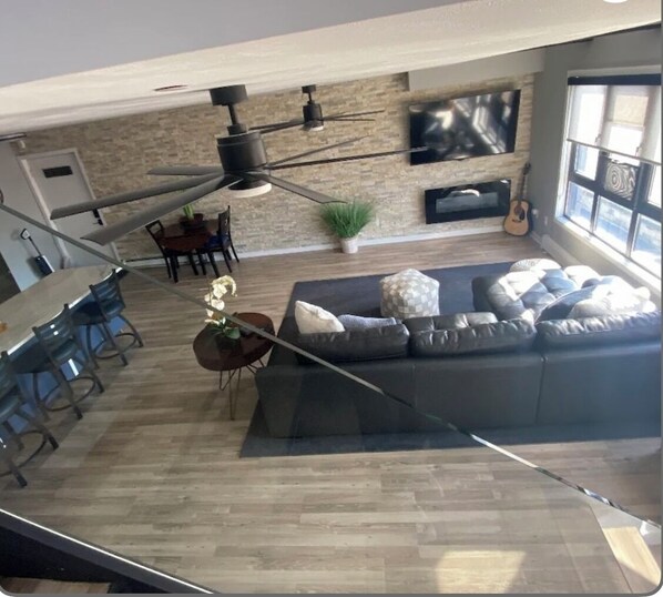 A modern living room with a large sofa, a dining area with chairs, a ceiling fan, a TV mounted on the wall, and a guitar.