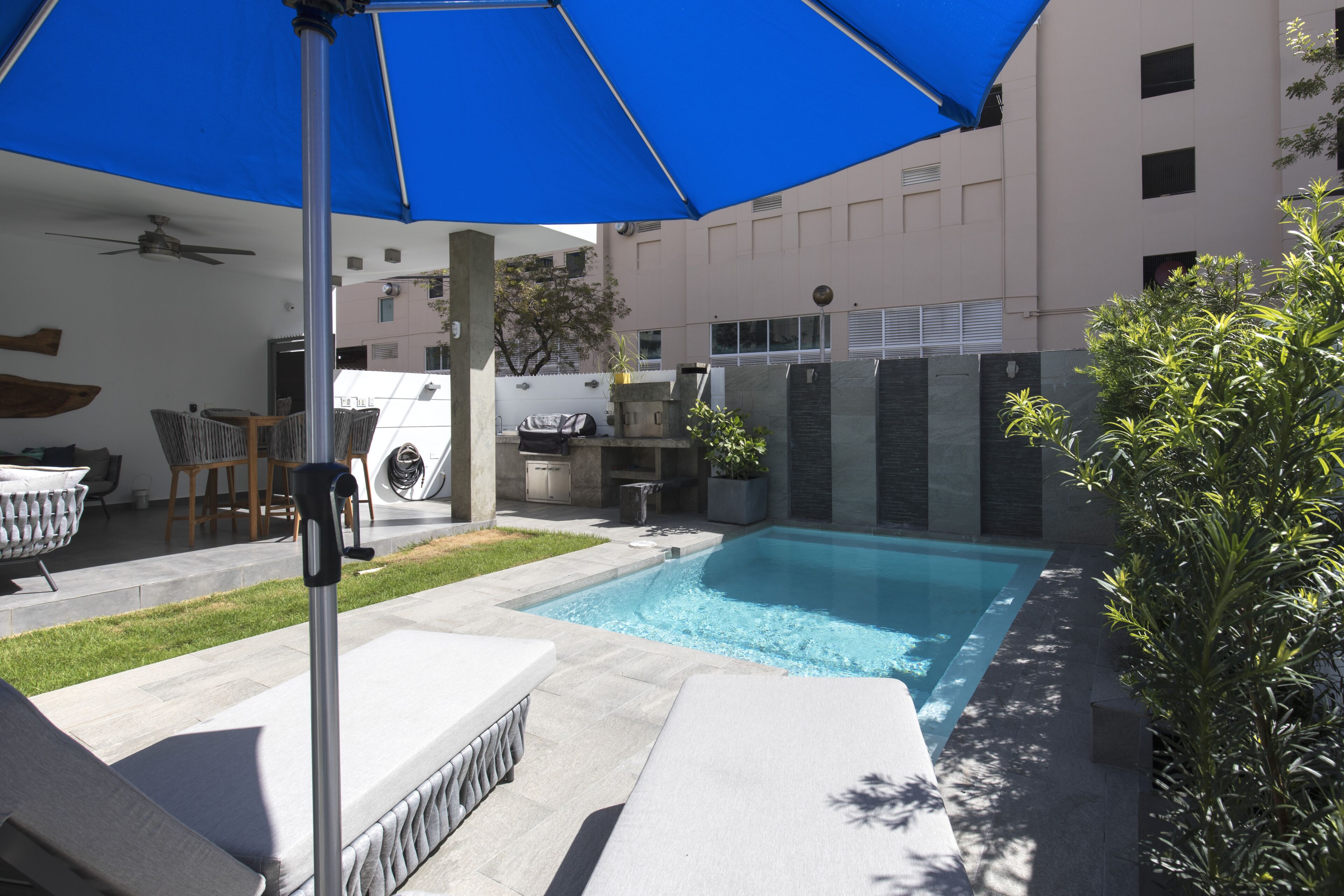 Un área de piscina moderna al aire libre con una sombrilla azul, sillas de descanso y un ventilador de techo.