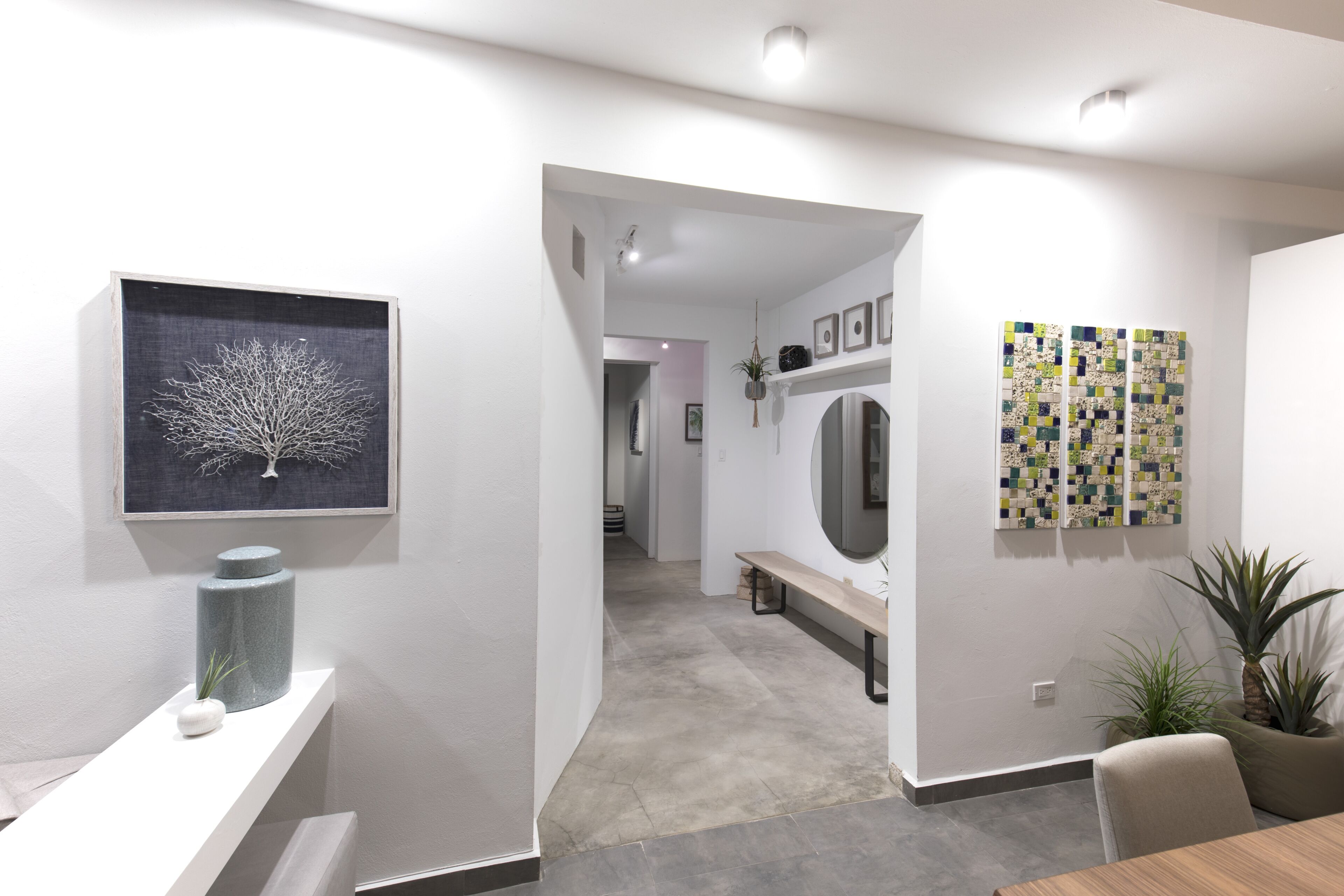 A modern interior with a hallway leading to a dining area, featuring a stone vase, a framed tree artwork, and a quilted wall panel.