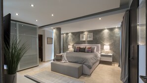 A modern bedroom with a large bed, bedside tables, a grey bench, and a glass partition.