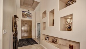Un couloir avec une porte en bois, un lustre et des étagères décoratives où sont posés divers objets, comme un vase et un cadre photo.
