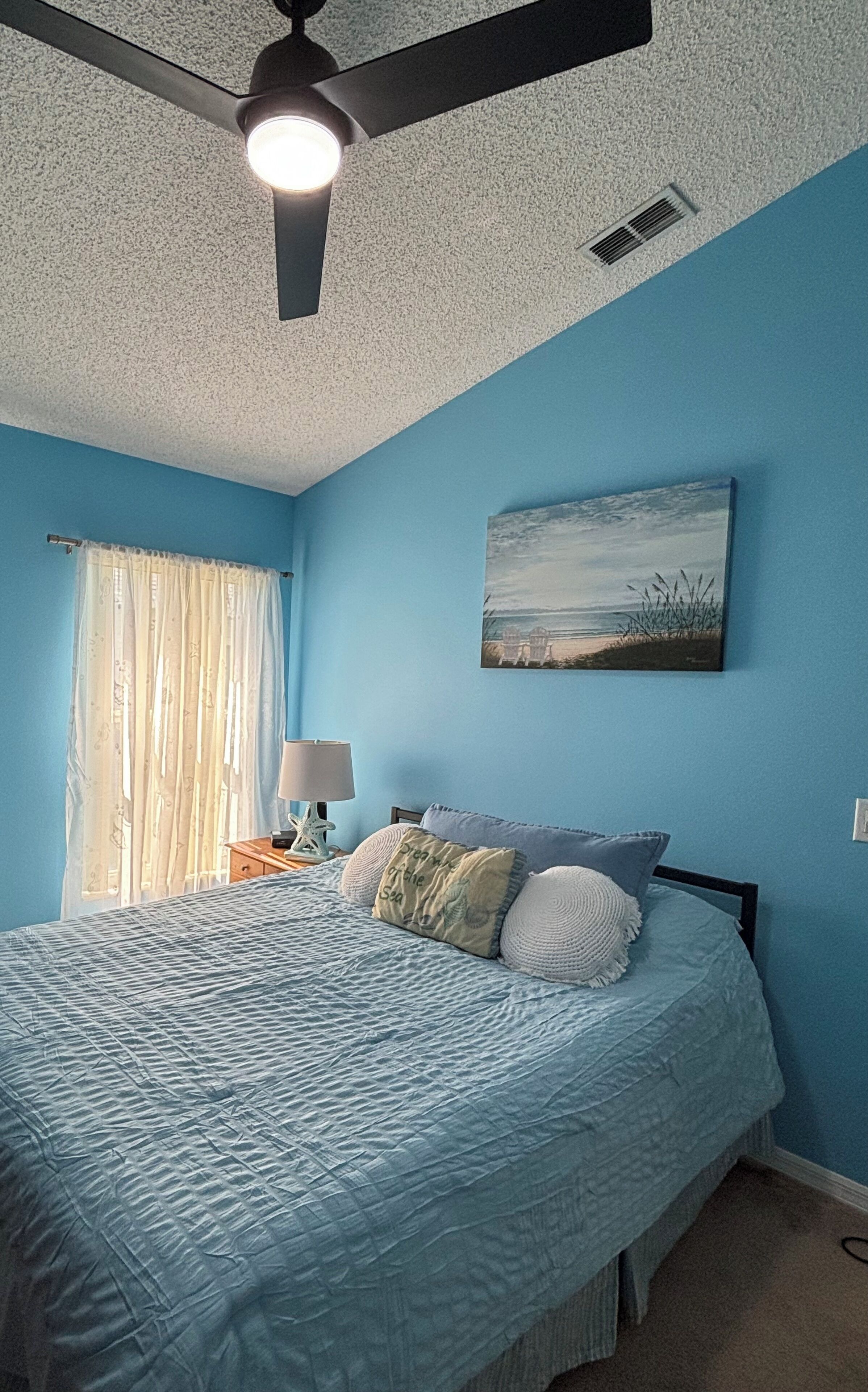 A bedroom with a bed, a ceiling fan, a window with curtains, and a painting on the wall.