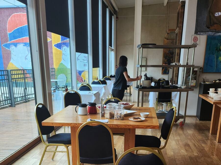 Restaurante com mesas e cadeiras de madeira, uma pessoa atendendo e um mural colorido na parede.