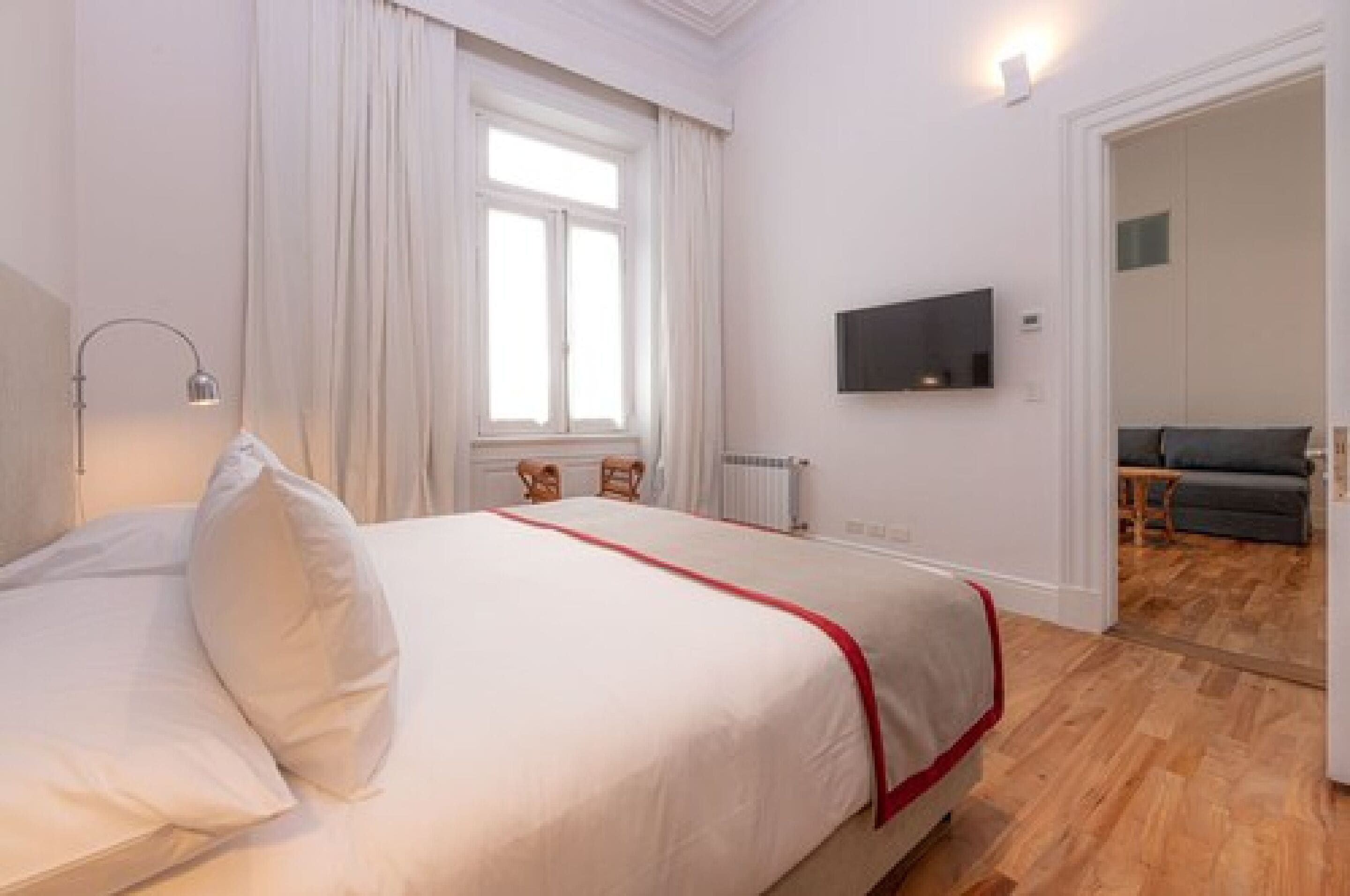 A hotel room with a bed, a TV mounted on the wall, a window with curtains, and a wooden floor.