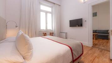 A hotel room with a bed, a TV mounted on the wall, a window with curtains, and a wooden floor.