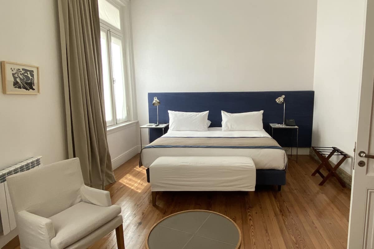 Habitación de hotel con cama, una silla, mesita de noche, lámpara y ventana con cortinas.