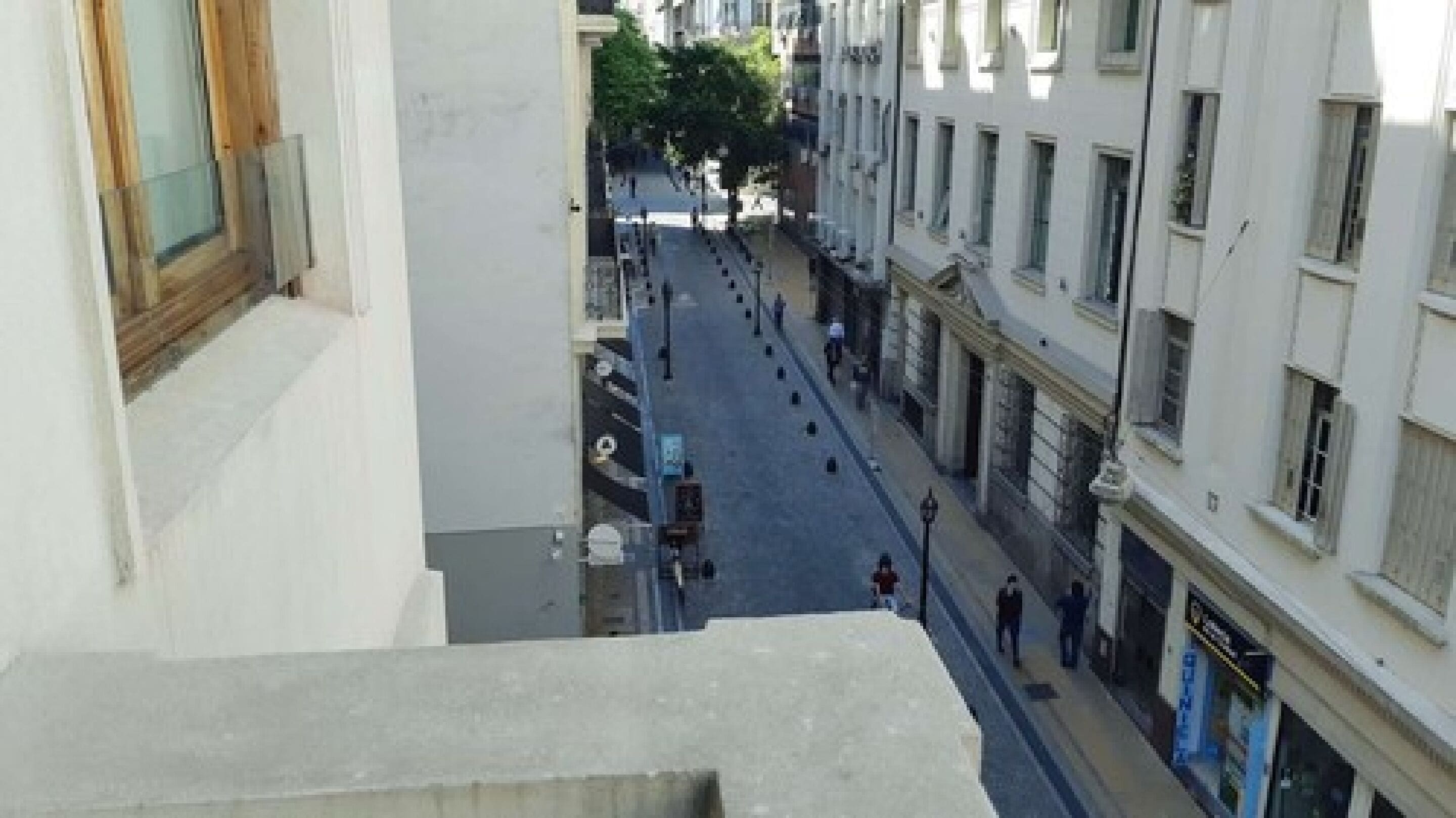 A narrow street with buildings on either side, a few pedestrians, and a tree in the middle.