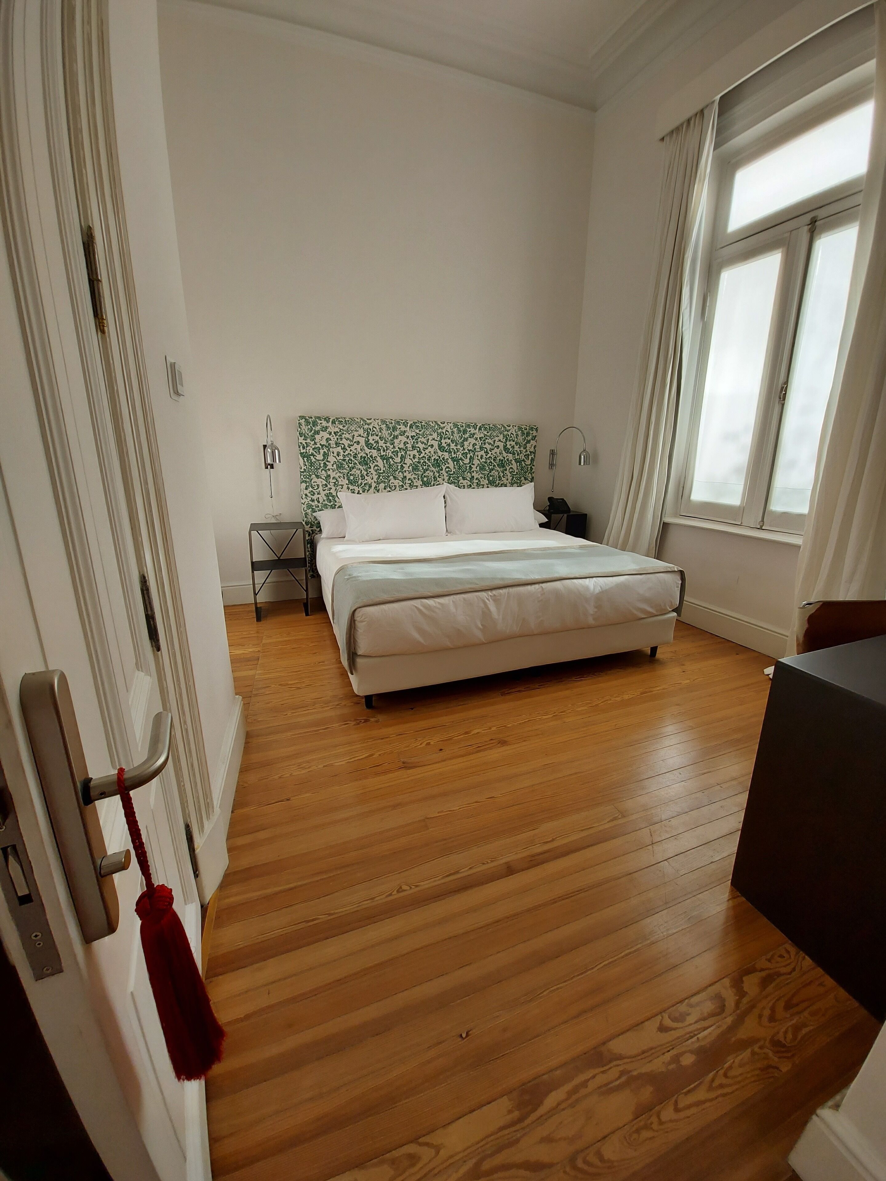 A bedroom with a bed, wooden floor, a nightstand, a lamp, and a window with curtains.