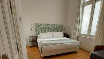 A bedroom with a bed, wooden floor, a nightstand, a lamp, and a window with curtains.