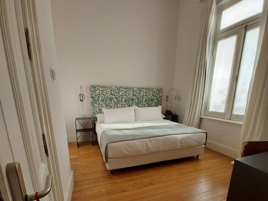 A bedroom with a bed, wooden floor, a nightstand, a lamp, and a window with curtains.