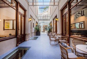 A covered outdoor café with wicker furniture and glass flooring, leading to a building with large windows and a sign reading "ESU". - Cassa Lepage Art Hotel Buenos Aires (Buenos Aires)