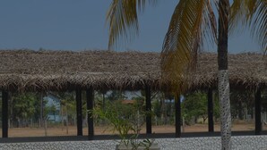 A covered outdoor area with a thatched roof, surrounded by palm trees and a stone wall. - Delight Guest House And Restaurant (Pasikuda)