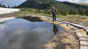 A person standing on a wooden boardwalk beside a reflective water body, with a mountainous landscape in the background.