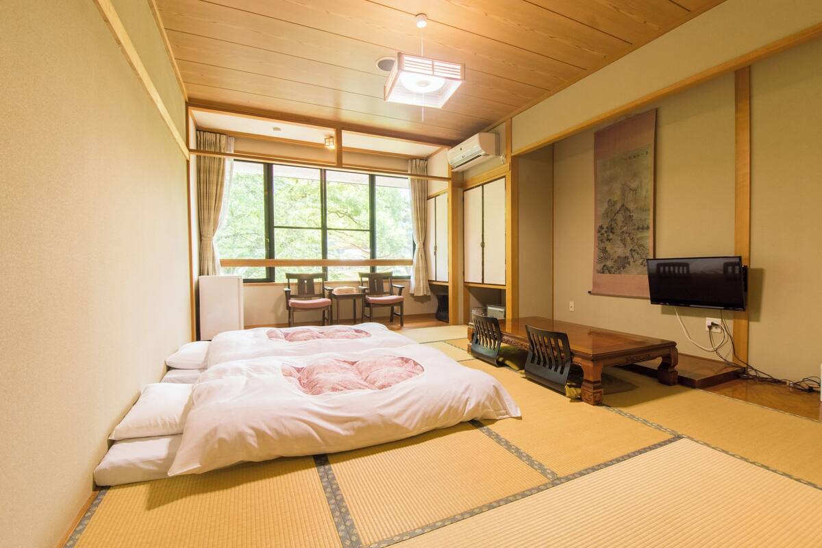 Une pièce de style japonais traditionnel, avec un sol recouvert de tatamis, un lit avec une literie blanche, une télévision et une porte coulissante donnant sur un balcon.