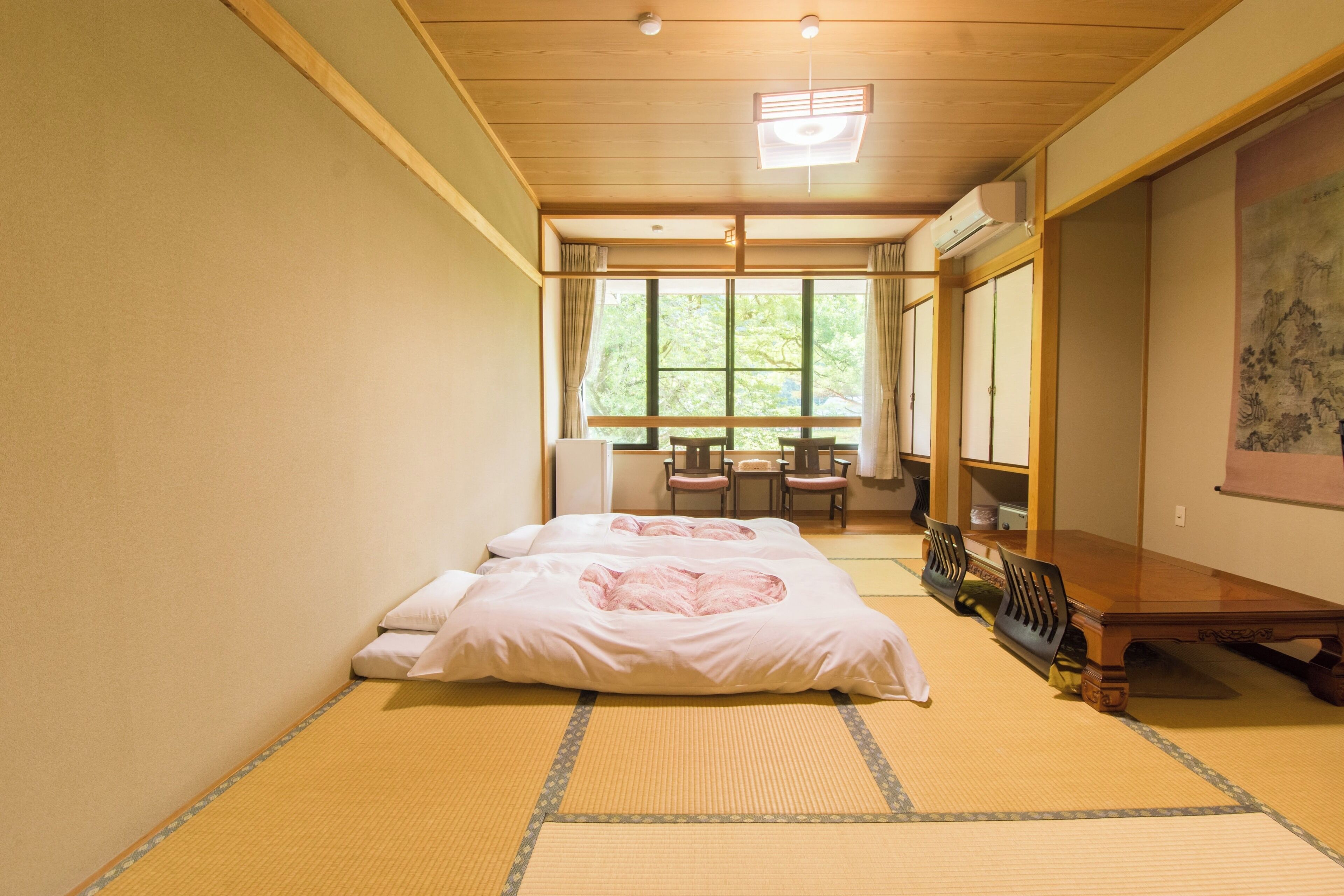 A traditional Japanese-style room with tatami flooring, a low wooden bed, a wooden table with chairs, and a large window with curtains.
