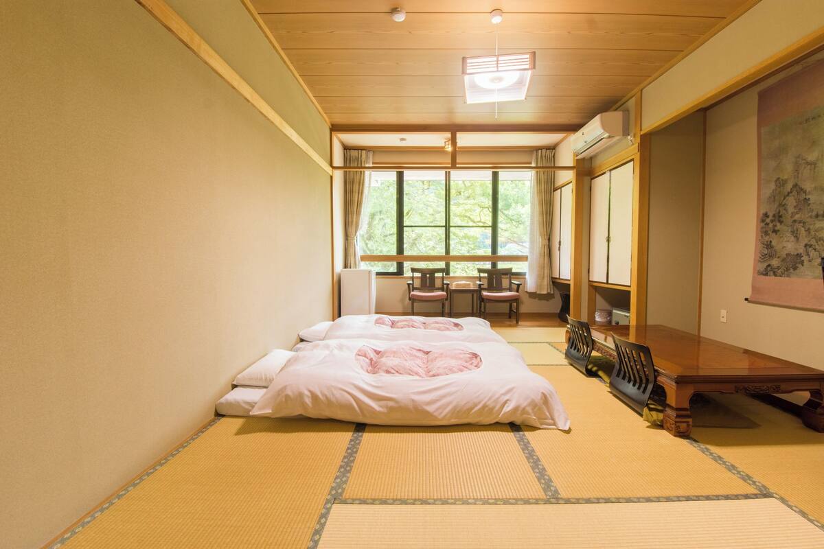 Une pièce de style japonais traditionnel, avec un sol recouvert de tatamis, un lit bas en bois, une table en bois entourée de chaises et une grande fenêtre avec des rideaux.