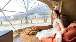A tent with a large window, a bed with orange bedding, and a view of a landscape with trees and buildings.