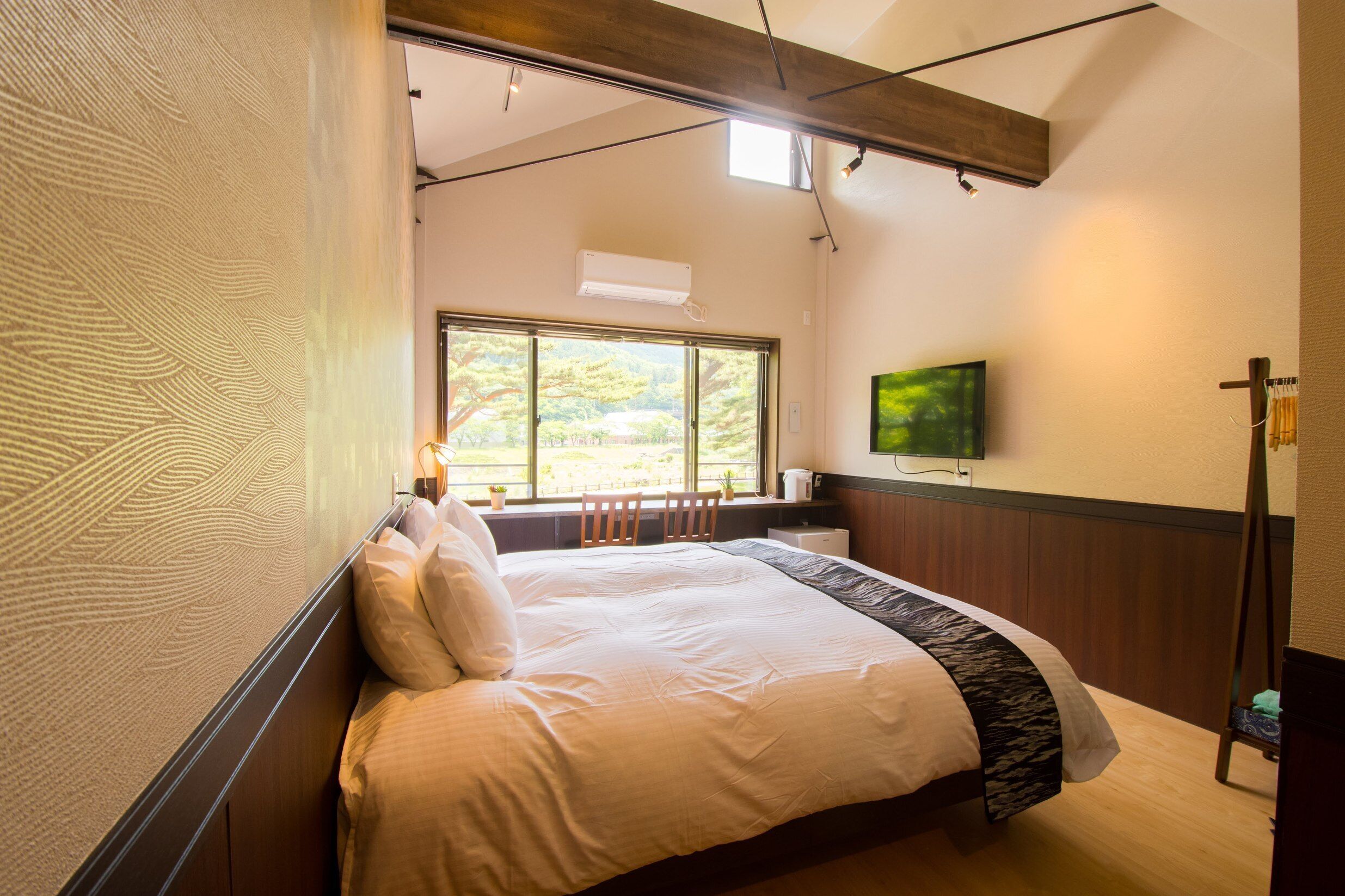 A bedroom with a bed, a TV mounted on the wall, a window, and a wooden ceiling beam.