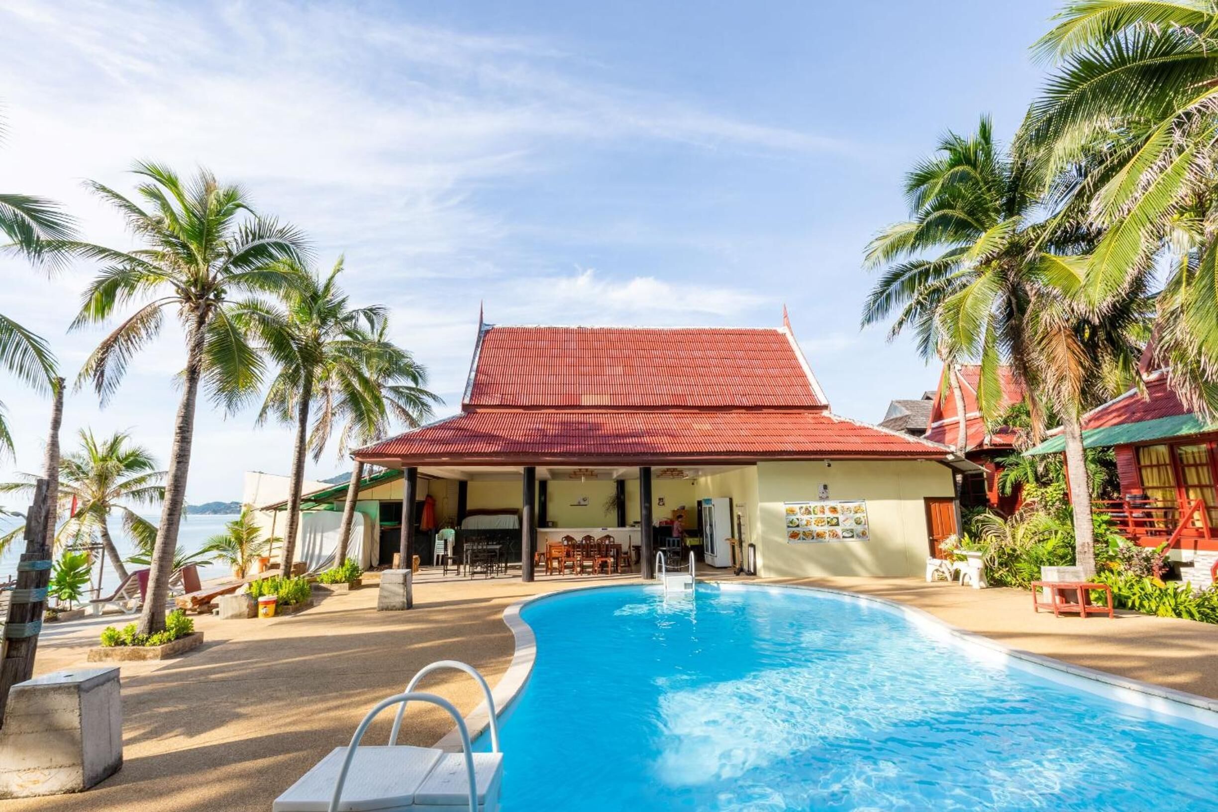 A poolside area with a covered seating area, palm trees, and a building with a red roof.
