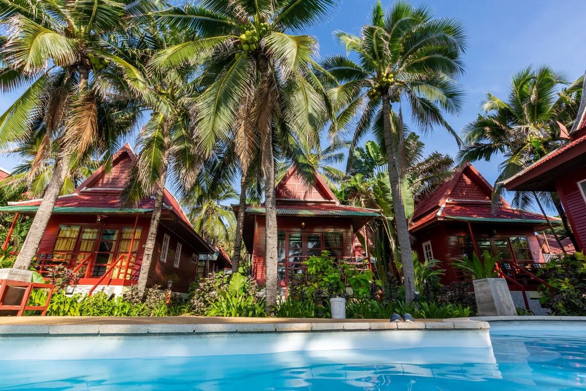A poolside view of a resort with red-roofed buildings, palm trees, and a clear blue sky.