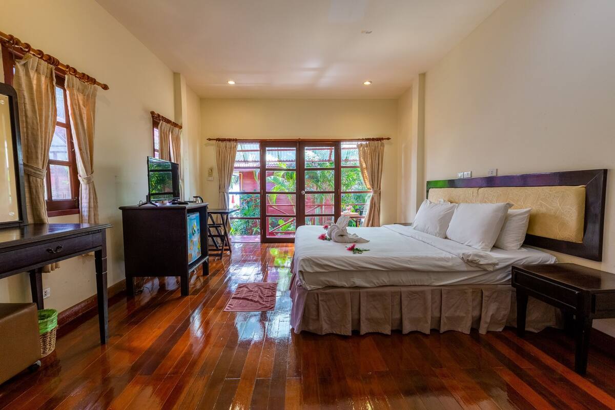 A hotel room with a wooden floor, a bed with white linens, a desk with a computer, and a view of greenery through the windows.