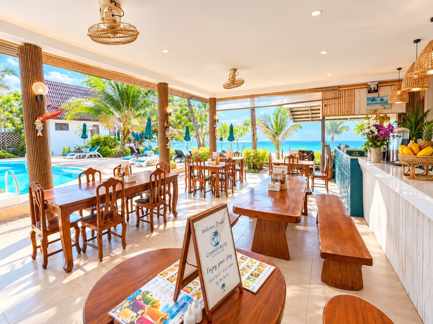 A beachfront restaurant with wooden tables and chairs, a pool, and a view of the ocean.
