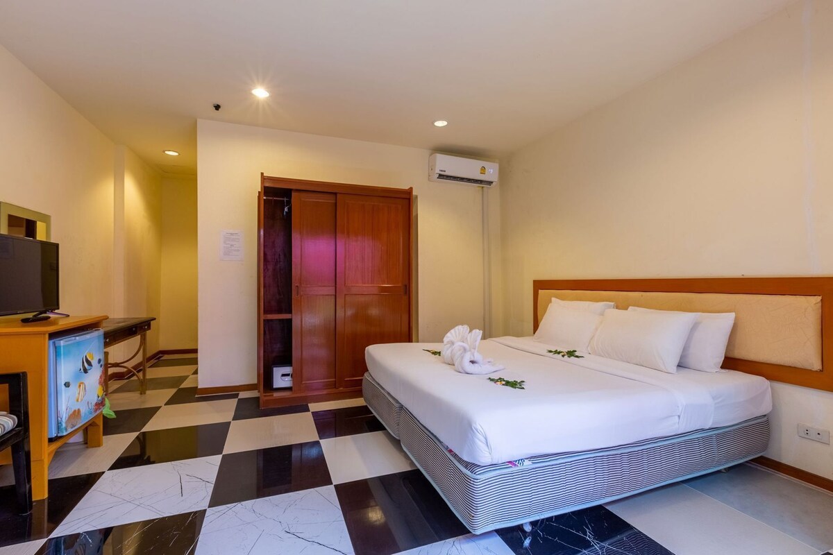 Una habitación de hotel con piso a cuadros, una cama con sábanas blancas, un cabecero de madera, un escritorio con televisor y un armario.