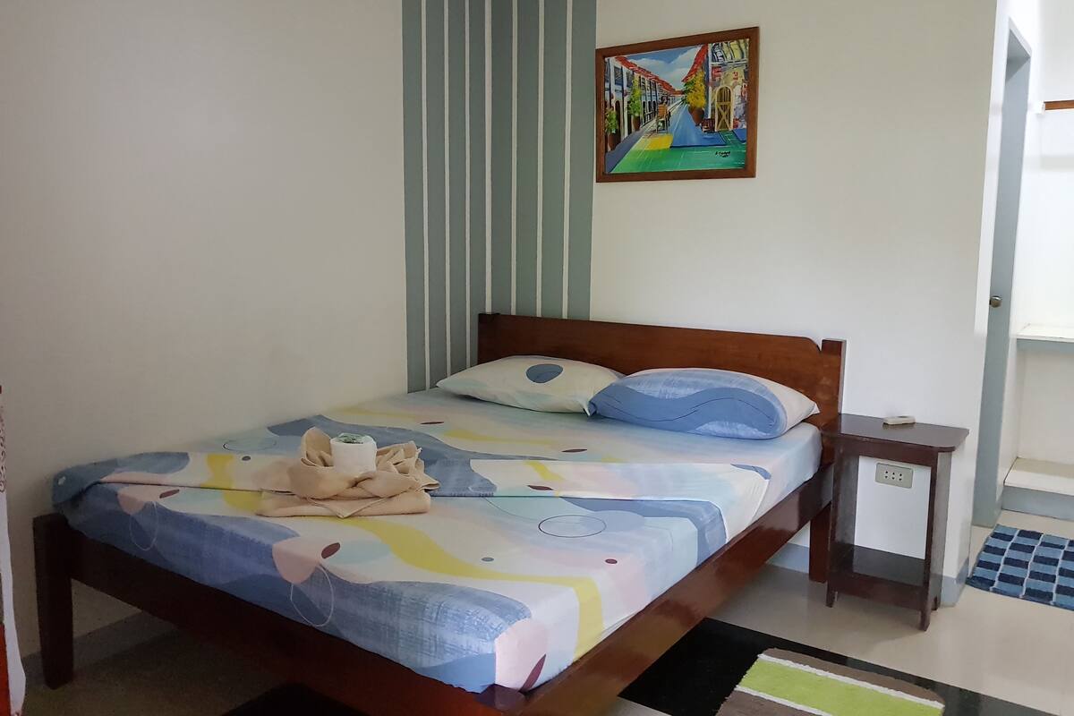 A neatly made bed with a colorful bedspread, a small bedside table with a lamp, and a painting on the wall.