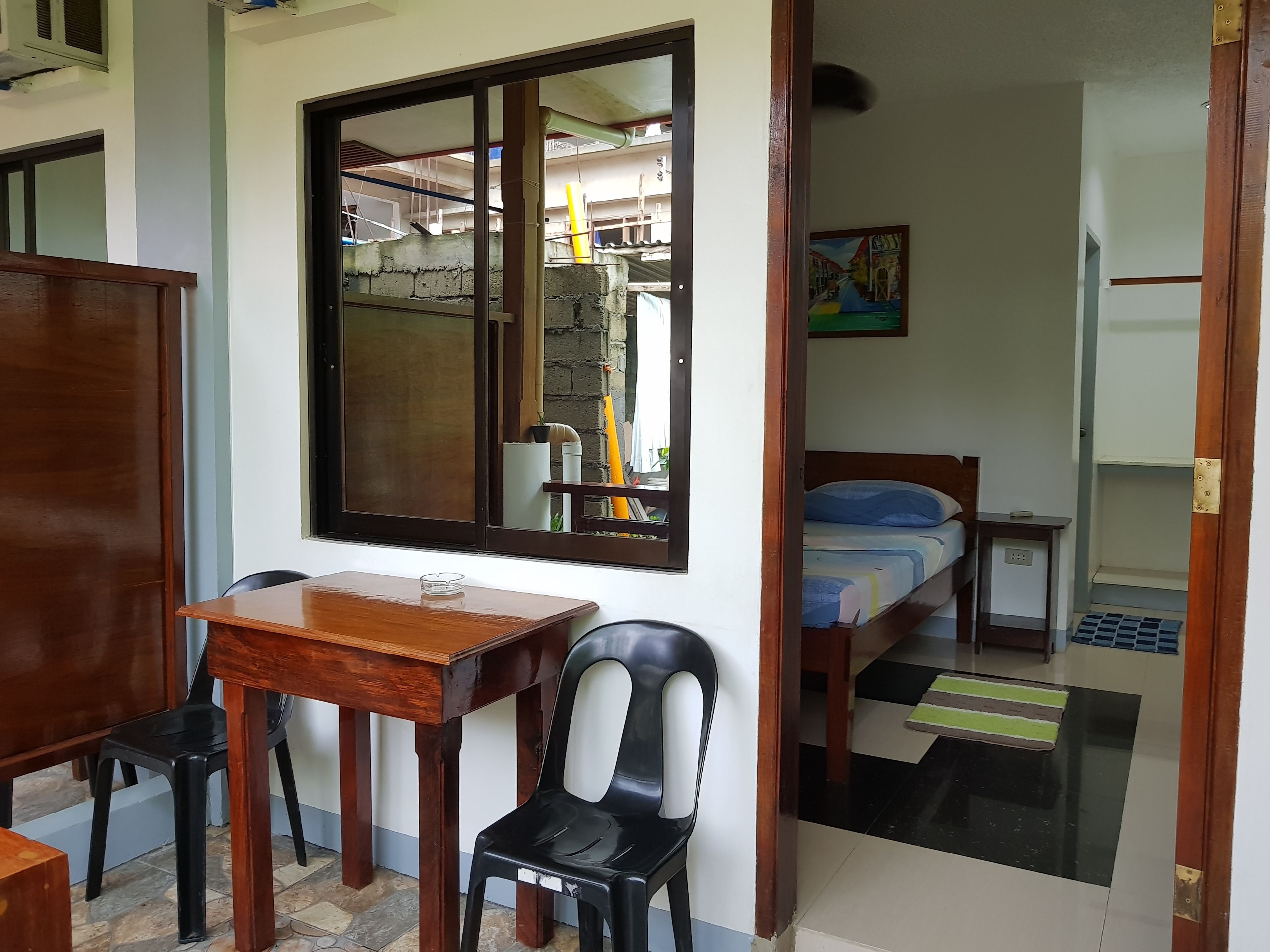 A room with a wooden table and chairs, a bed with a blue mattress, a small bedside table, and a window with a view of the outside.