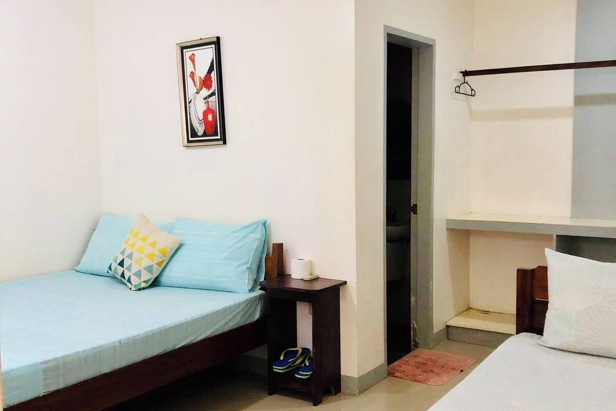 A bedroom with two beds, a ceiling fan, a framed picture, and a small table with slippers.