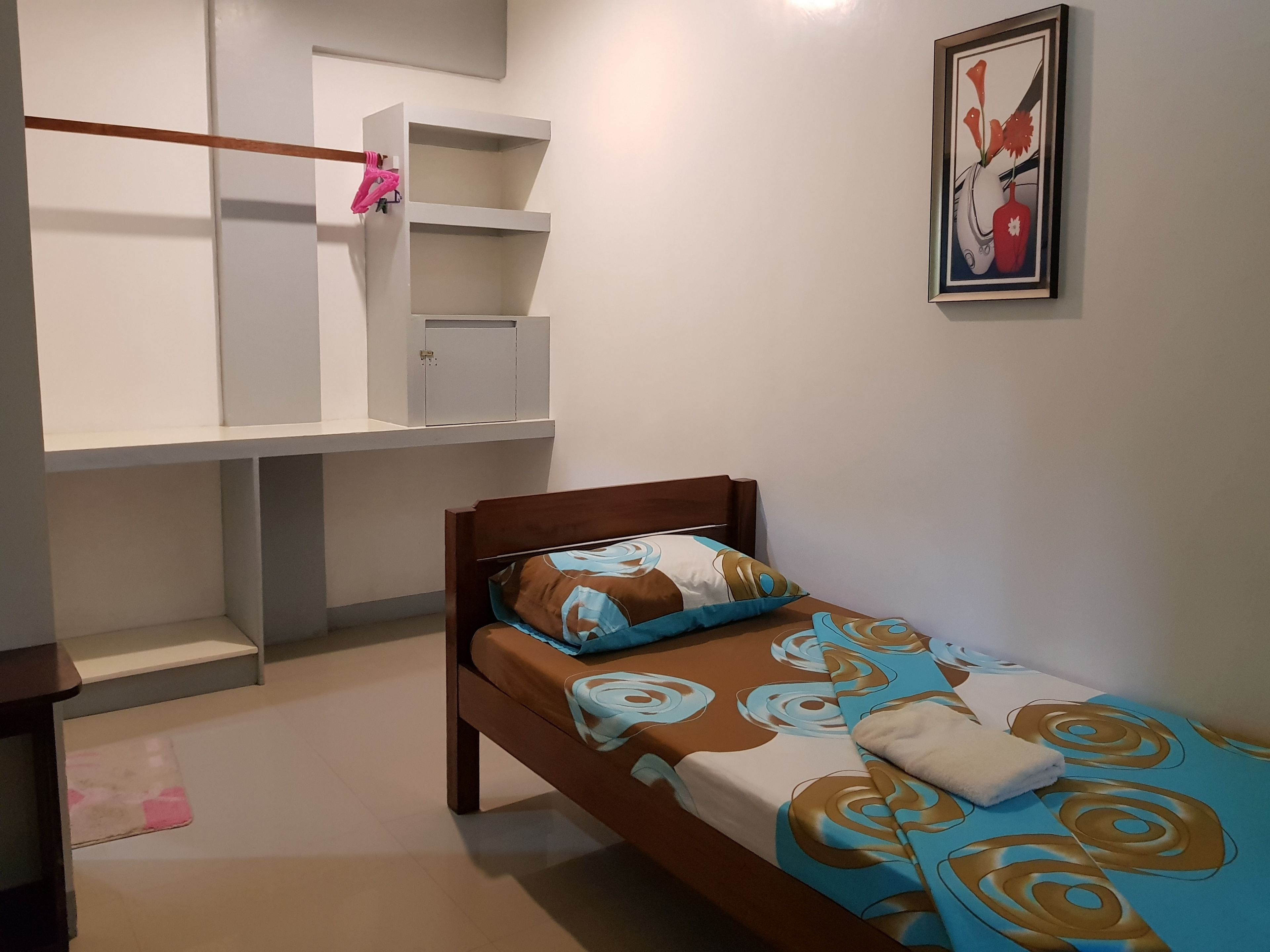 A small, neatly organized bedroom with a single bed, a wardrobe, and a framed artwork on the wall.