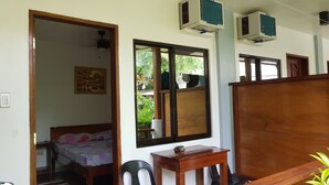 A room with a bed, a desk, two air conditioning units, a mirror, and a window with a view of greenery. - Hidden Garden Pension (El Nido)