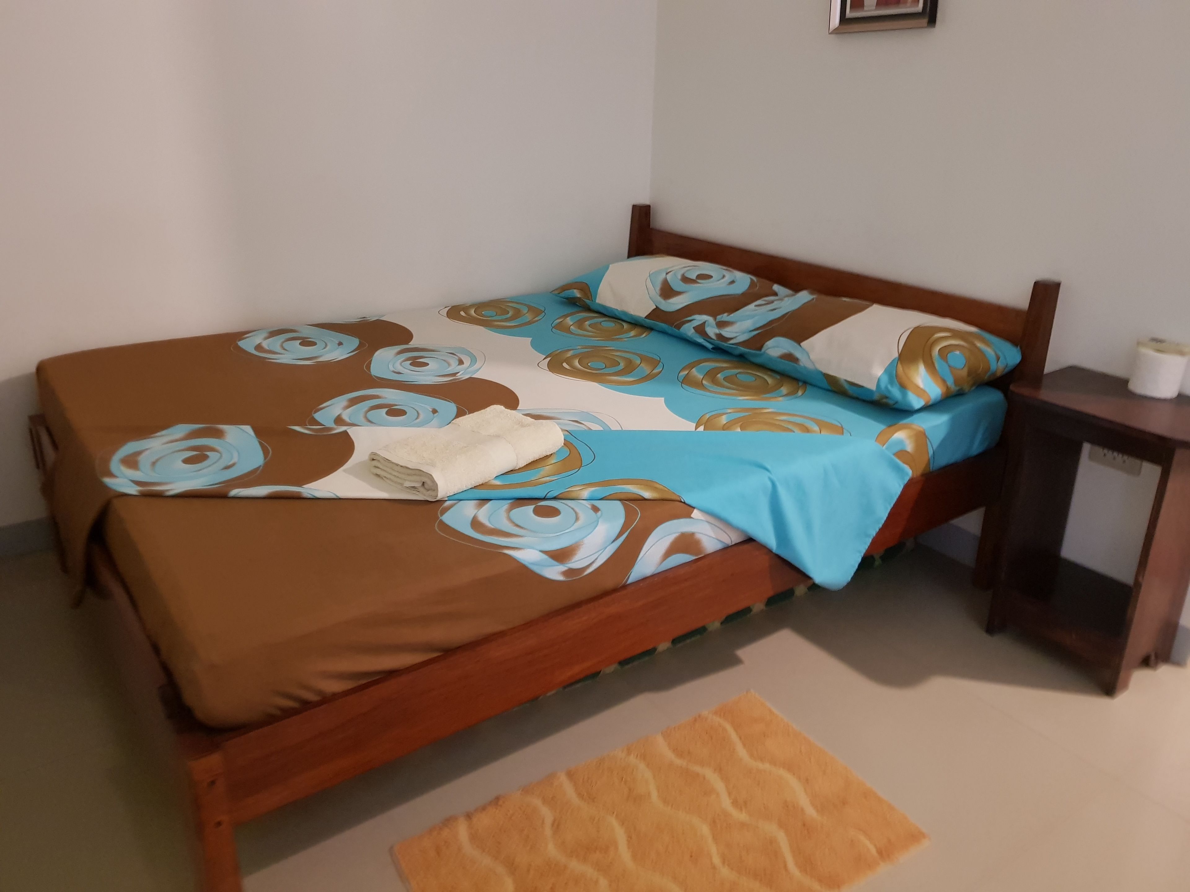 A bed with a brown and blue floral bedspread, a folded white towel, and a wooden headboard.