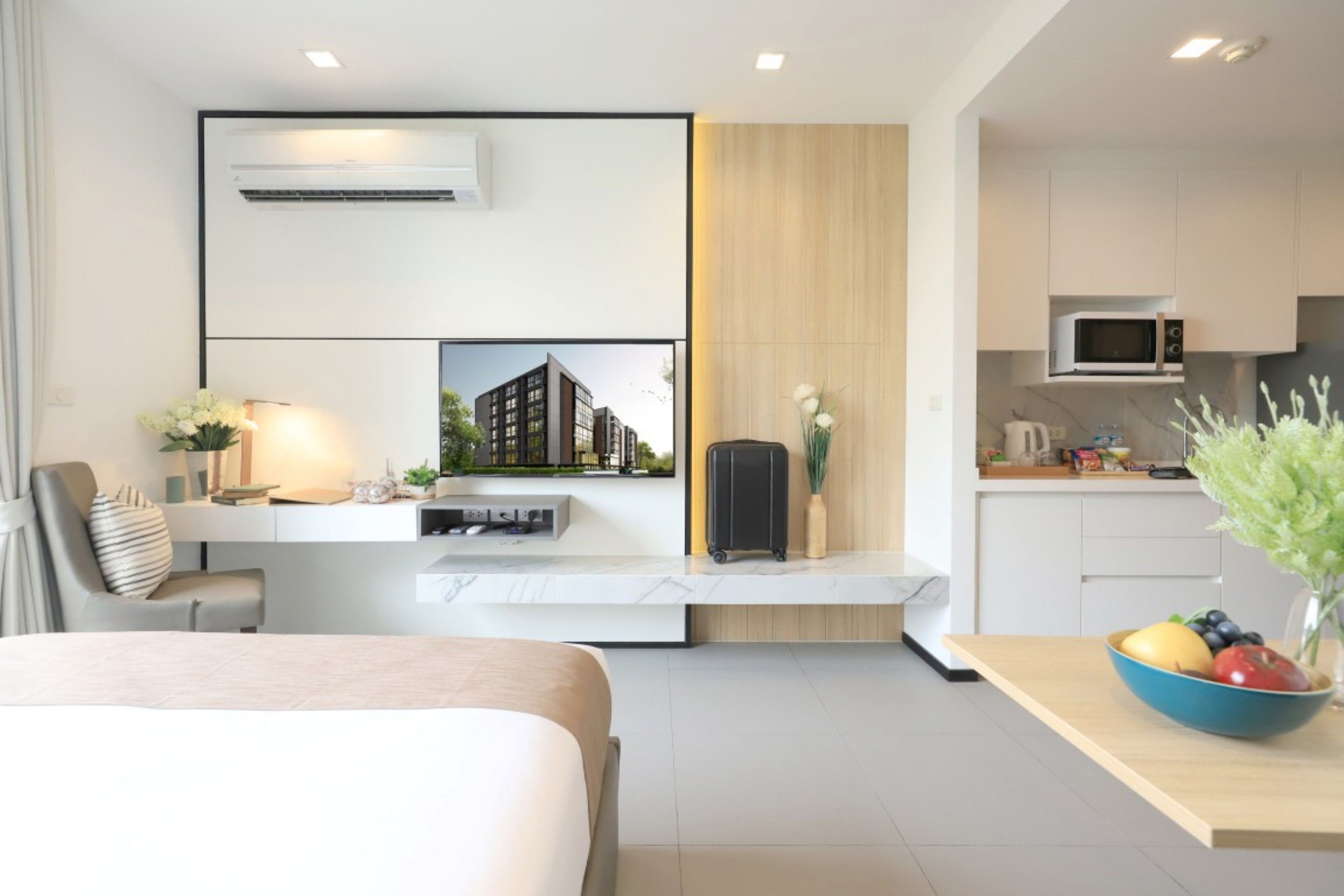A modern hotel room with a bed, a desk with a lamp, a small kitchenette, and a view of a building.