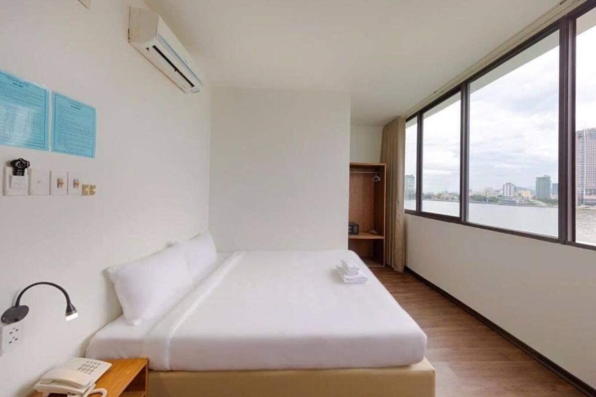 A hotel room with a bed, a bedside telephone, a window with a city view, and a wall-mounted air conditioner.