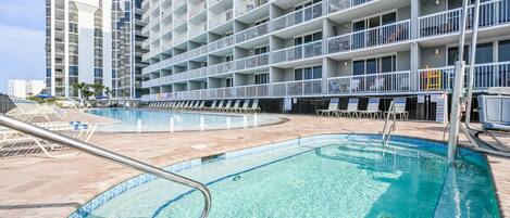A hotel pool area with clear blue water, lounge chairs, and a multi-story building with balconies.