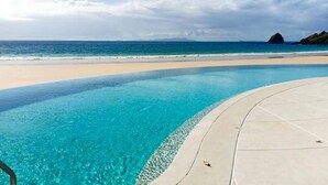 Una piscina azul cristalina con borde curvo, una playa arenosa y vistas al océano y formaciones terrestres distantes.