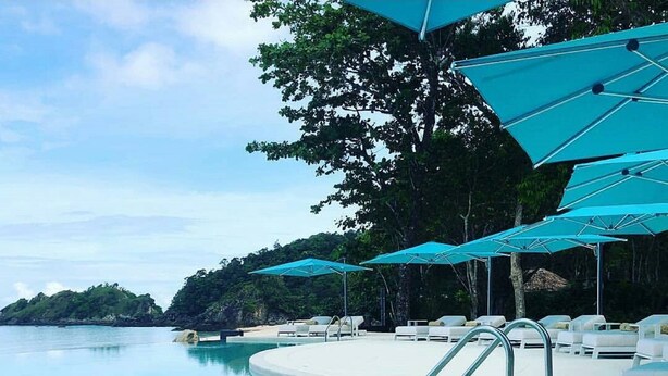A poolside area with lounge chairs and umbrellas, surrounded by trees and overlooking a body of water.