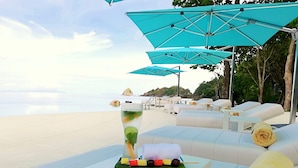A beachside lounge area with white cushioned seating, a glass of iced beverage, and a skewer of colorful fruit.