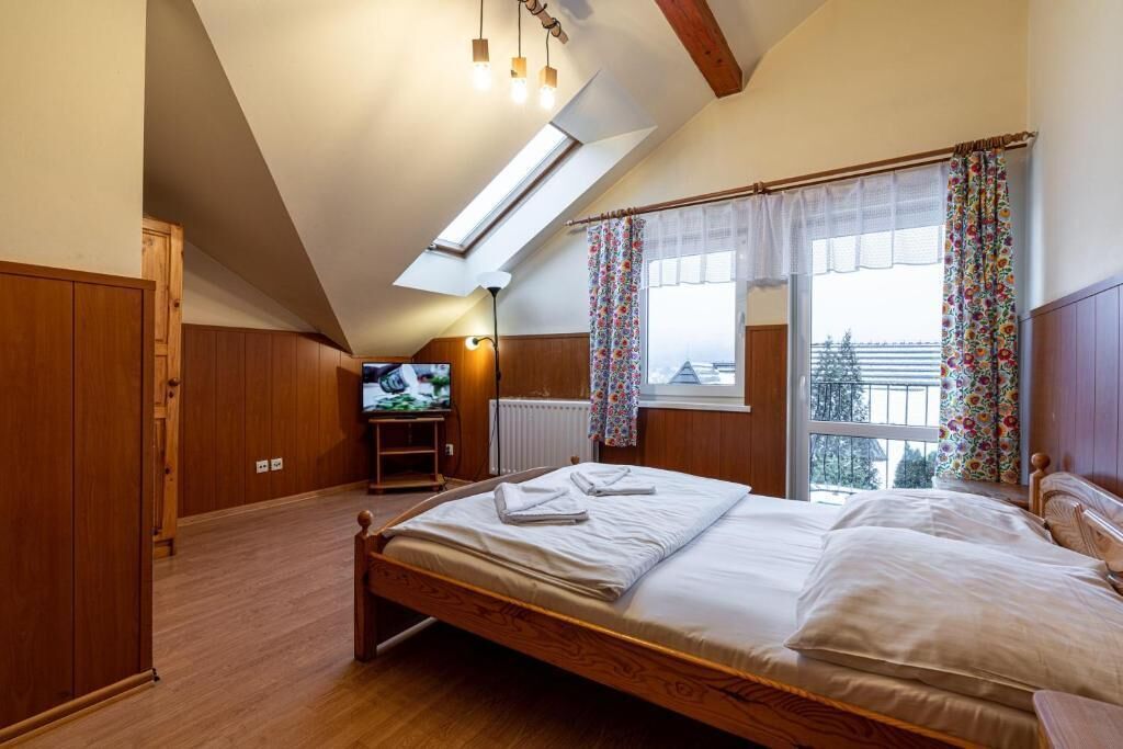 A bedroom with a sloped ceiling, a bed, a television, and a window with curtains.