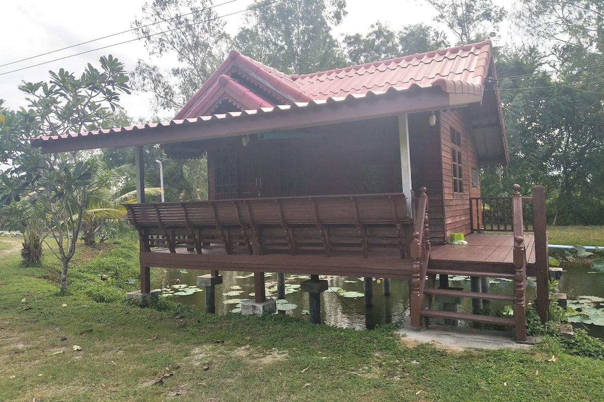 A wooden structure with a sloped roof, a porch with benches, and a pond with lily pads.
