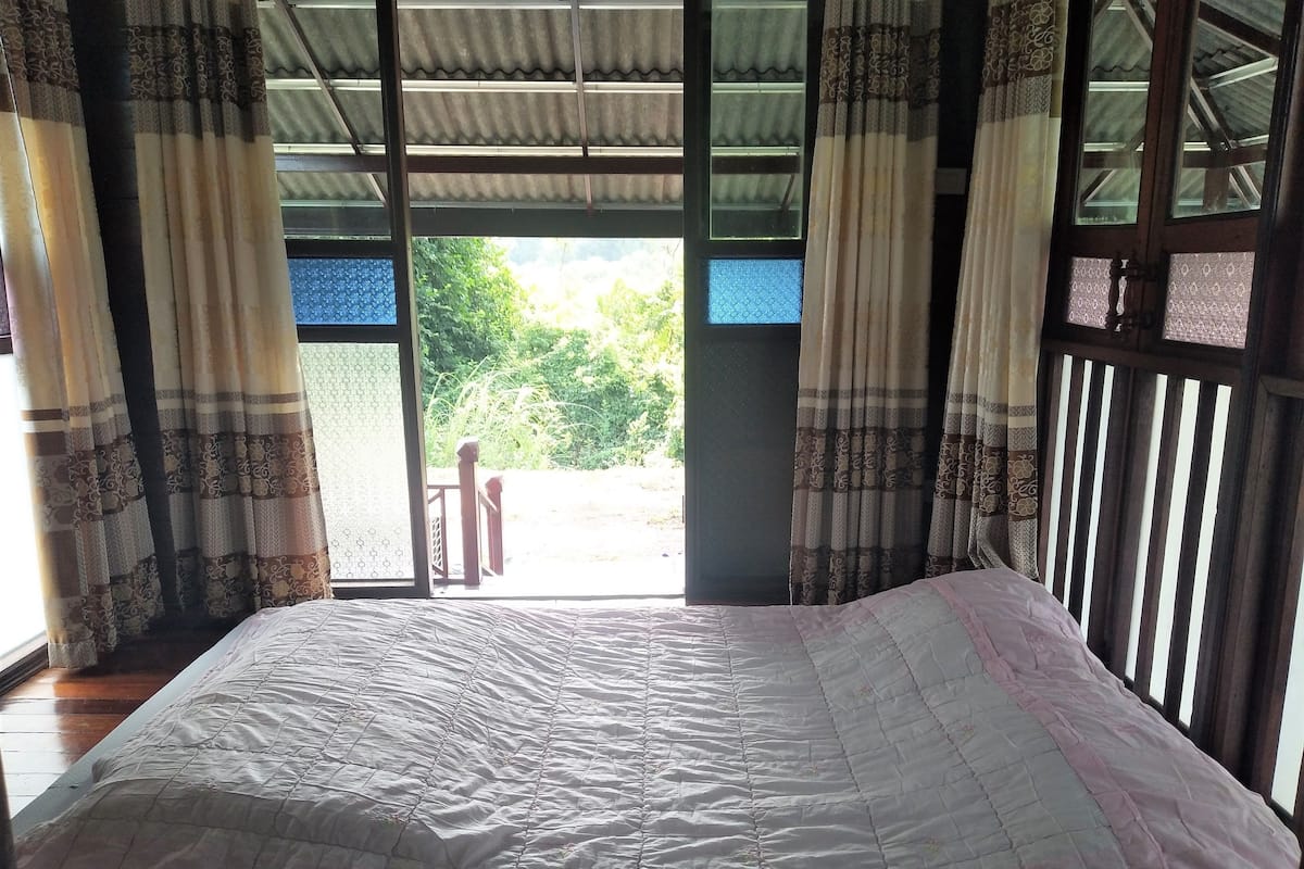 A bed with a quilt, curtains, and a view of greenery outside.