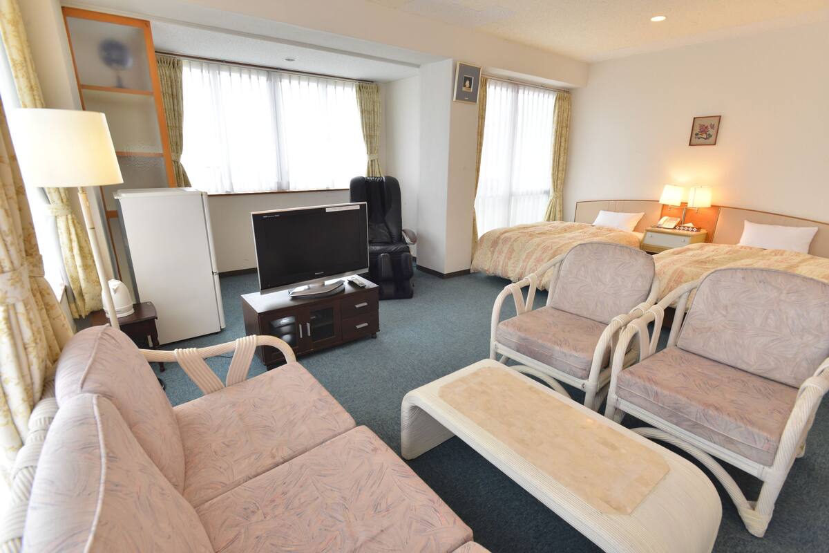 Ett hotellrum med en soffa, två fåtöljer, ett litet bord, en TV, ett minikylskåp, en lampa och ett sängbord med en lampa.
